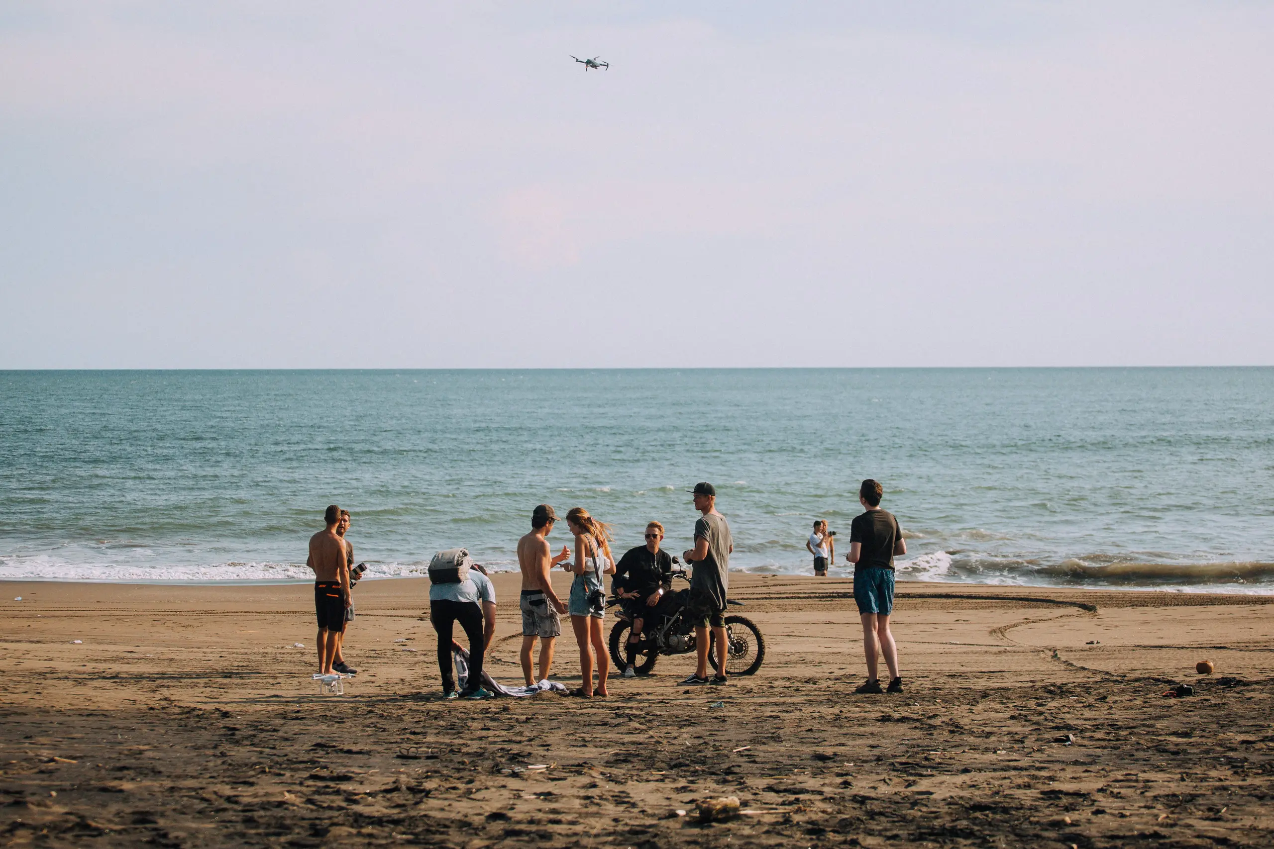 Sekelompok orang dan sebuah motor trail di pantai berpasir hitam, dengan laut dan langit sore di belakang