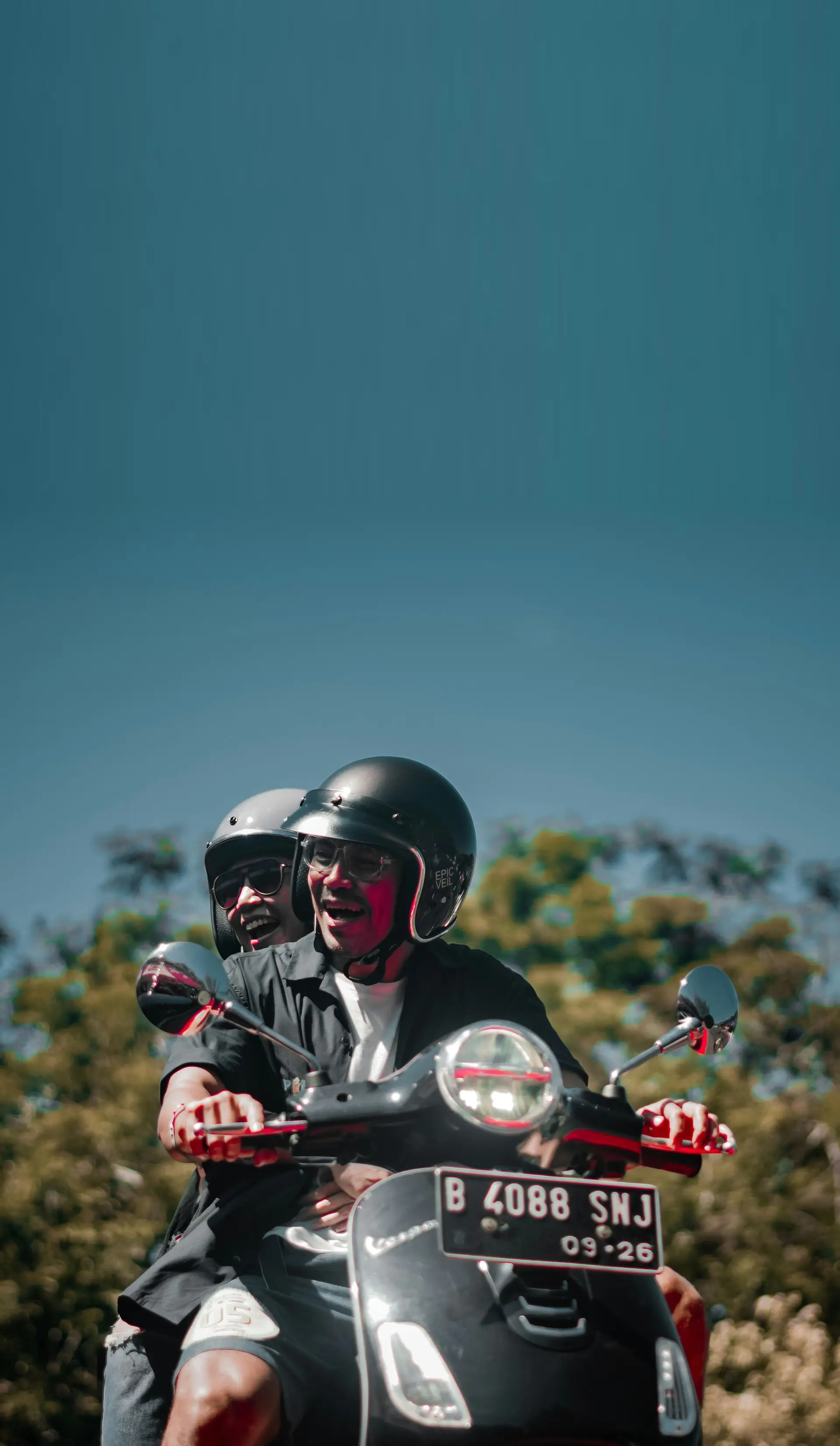 Dua orang berboncengan naik Vespa hitam sambil tertawa, berlatar pepohonan dan langit biru